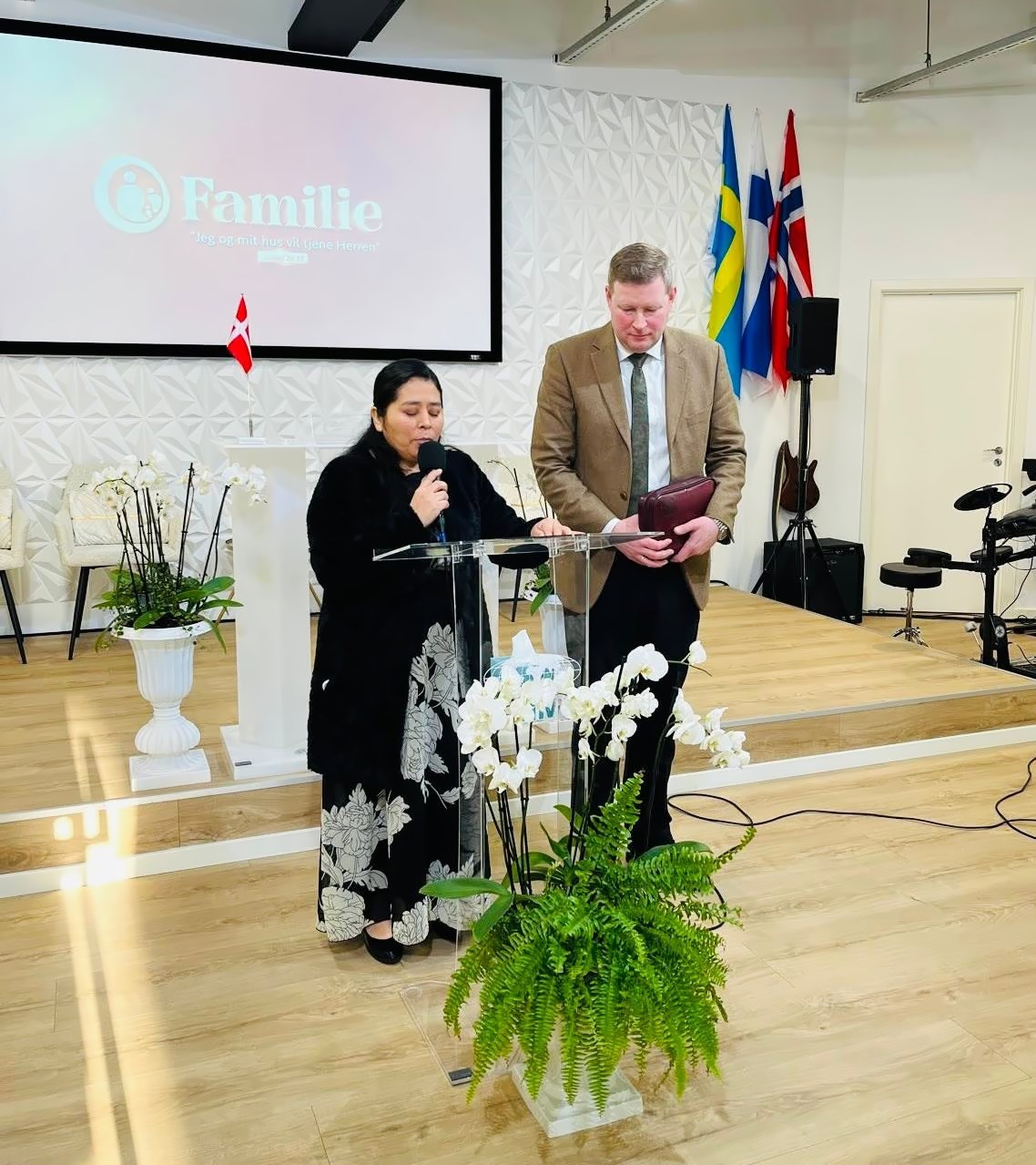 Una hermana habla al micrófono en el púlpito junto a un hermano con una Biblia; en la pantalla dice «Familie»