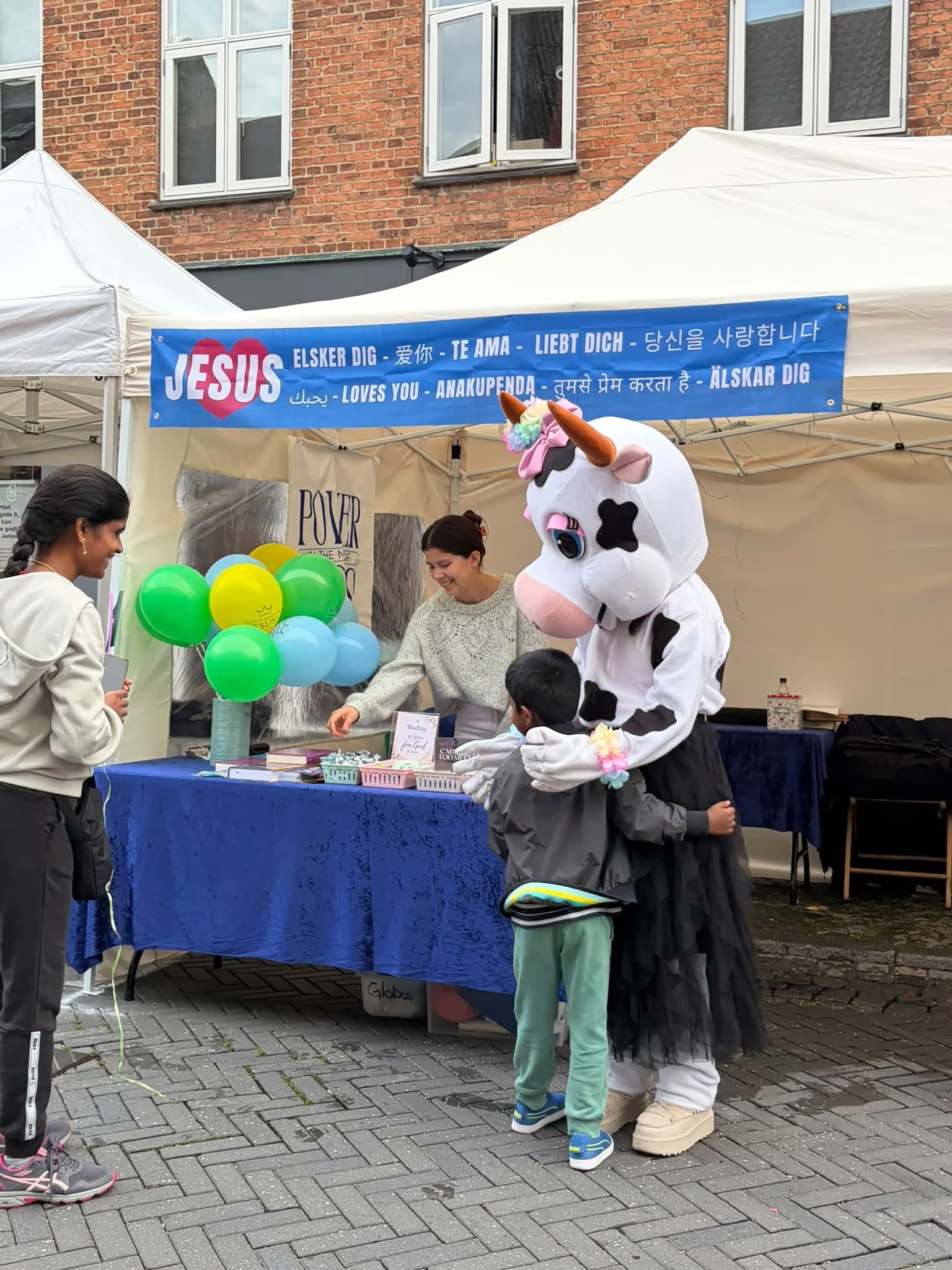 El puesto de la iglesia con globos y un cartel que dice «Jesús te ama» en varios idiomas; una mascota abraza a un niño